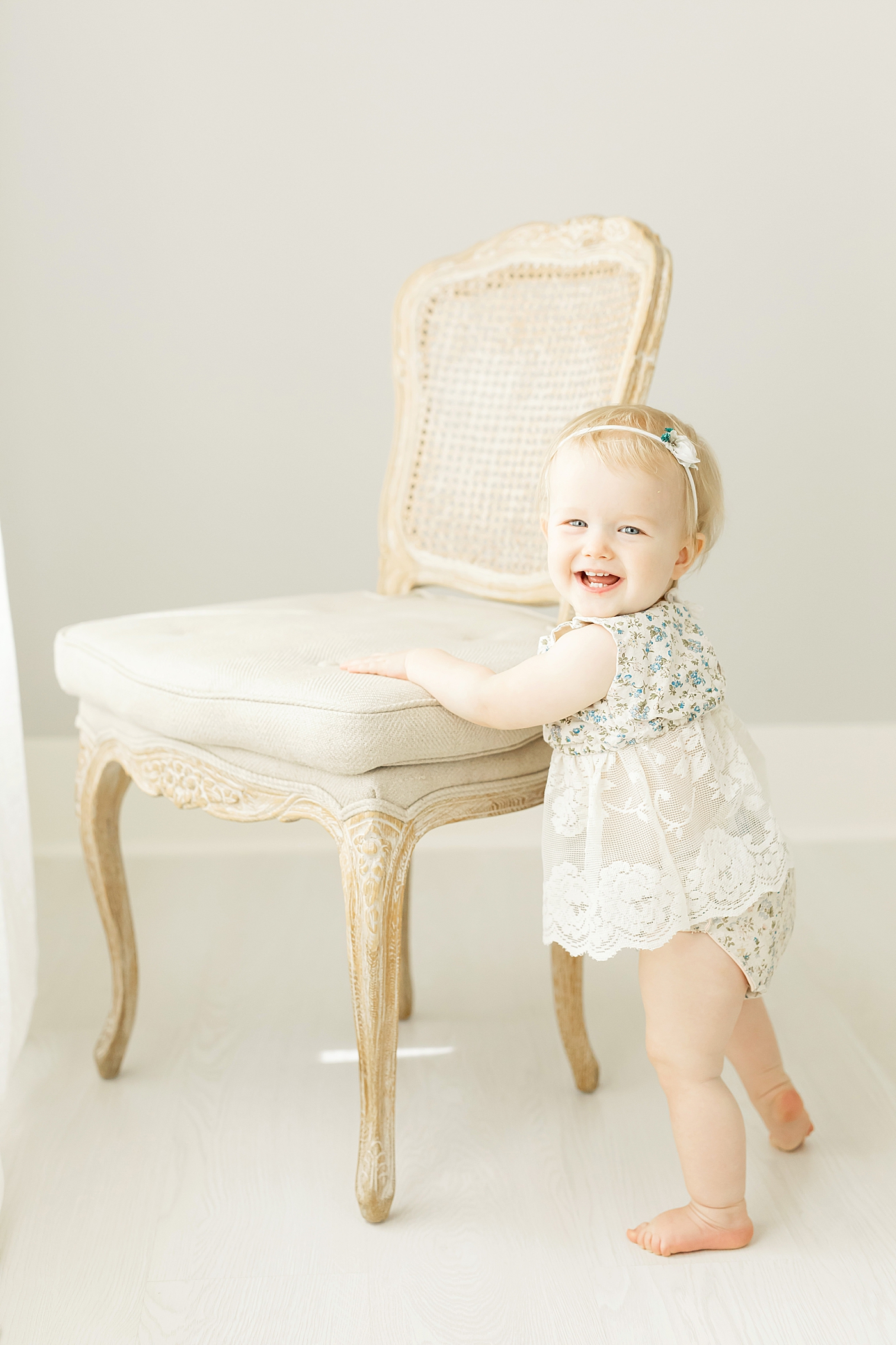 One year old in floral and lace romper standing up by a chair. Photo by Fresh Light Photography.