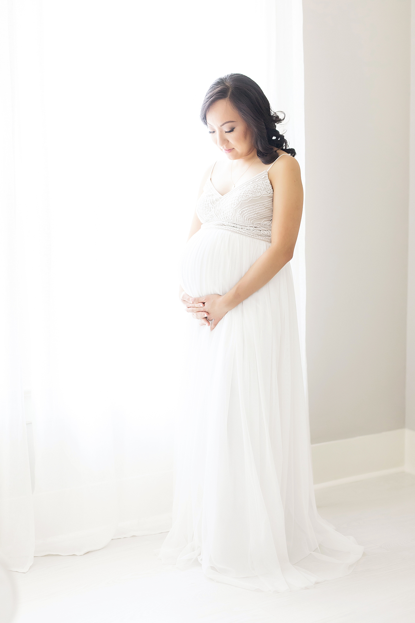 Pregnant mom in beautiful white dress standing in front of a window. Photo by Fresh Light Photography.