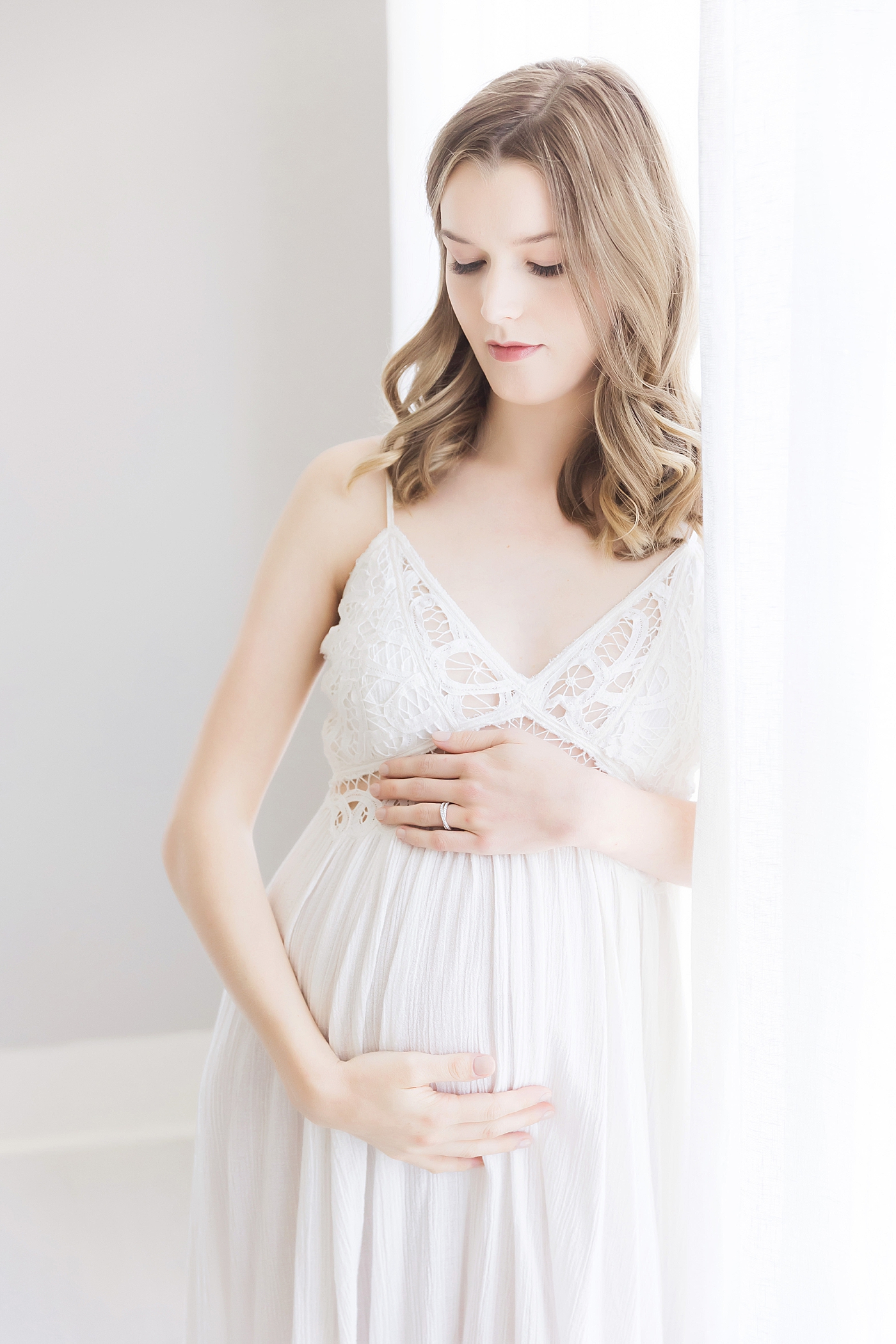 Expecting mom in white lace dress against window for maternity photos. Photo by Fresh Light Photography.